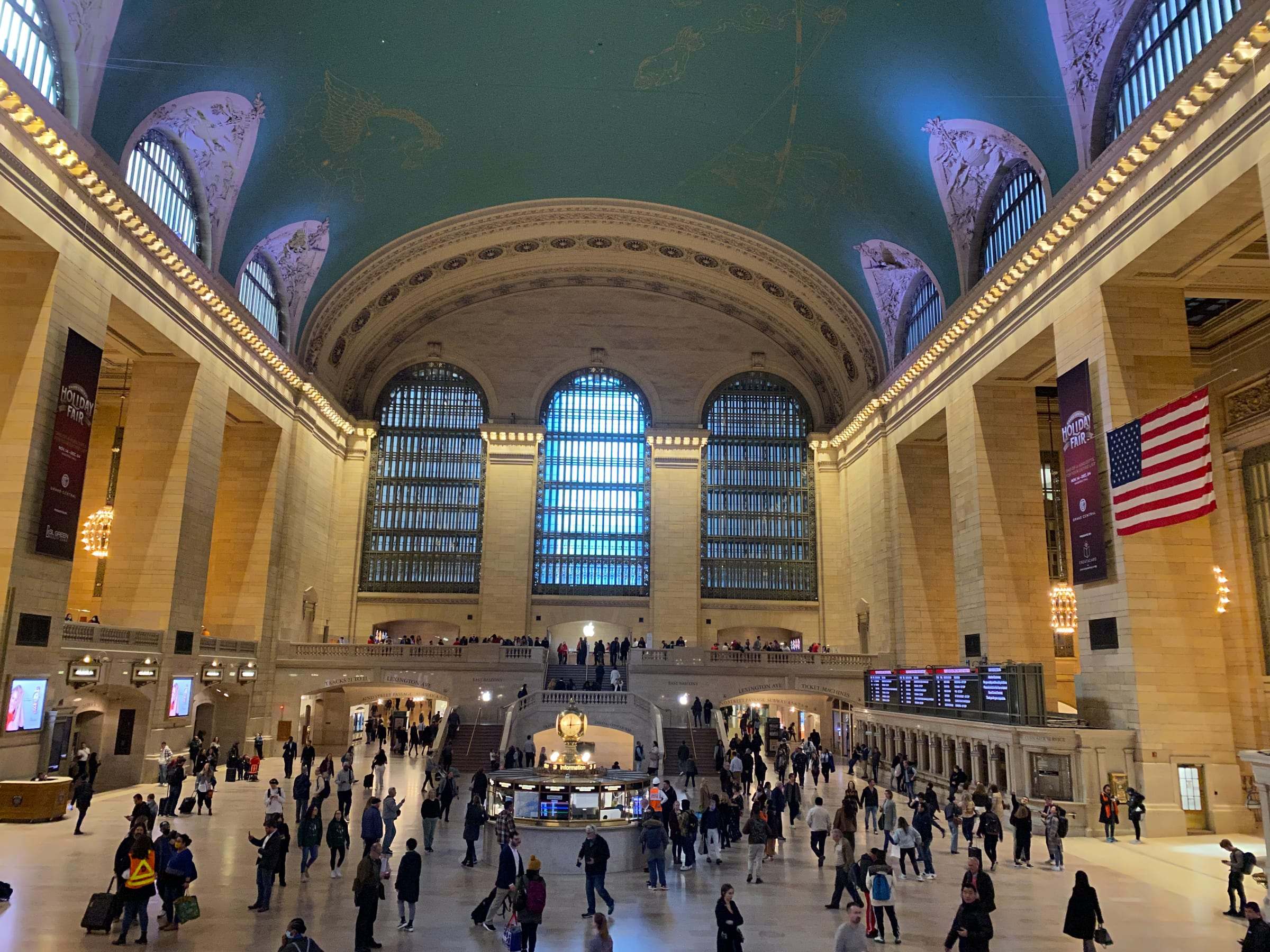Main concourse of Grand Central Terminal in New York with travelers, arched windows and an American flag